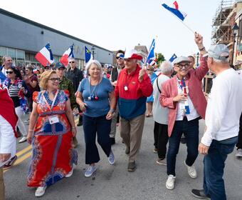 Governor General Mary Simon and Mr. Whit Fraser are walking with a crowd of people outside, participating in National Acadian Day festivities.