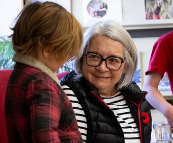  Governor General Mary Simon sits in a red chair, while holding a book and smiling at child who is standing beside her.