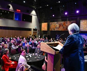 La gouverneure générale Mary Simon est vue de dos, debout, parlant à un podium, sur une scène, devant une salle remplie de personnes assises à des tables rondes.