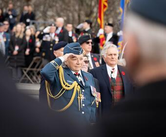 Governor General Mary Simon, wearing the Air Force uniform, is saluting. There is a crowd of people behind her. Her husband Whit Fraser is on her right.