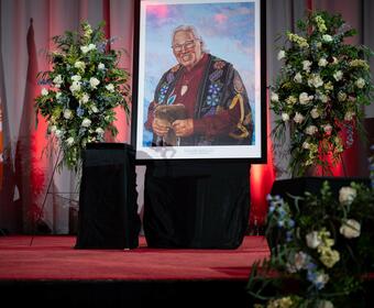 Painted portrait of Murray Sinclair showcased on a stage covered with red carpet ,surrounded by funeral flowers arrangements.