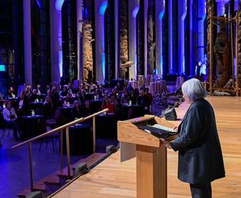 Governor General Mary Simon is seen from the left side, speaking at a podium, in front of a crowd of people sitting at round tables. Purple and lite blue lights are projected on tall pillars behind the audience.