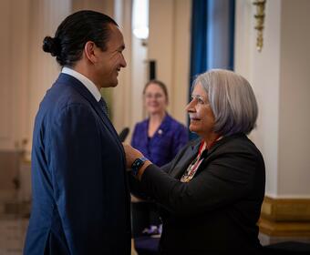 Governor General Simon is smiling to Premier Wab Kinew while pining a Coronation Medal on his vest.