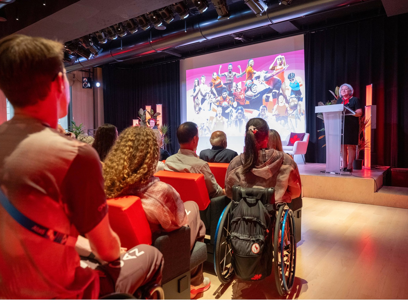 Un groupe de personnes dans un auditorium fait face à une scène où la gouverneure générale du Canada, Mary Simon, s'exprime depuis un podium. Derrière elle, un écran de projection affiche un collage d'images d'athlètes. Parmi le public, se trouvent des pe