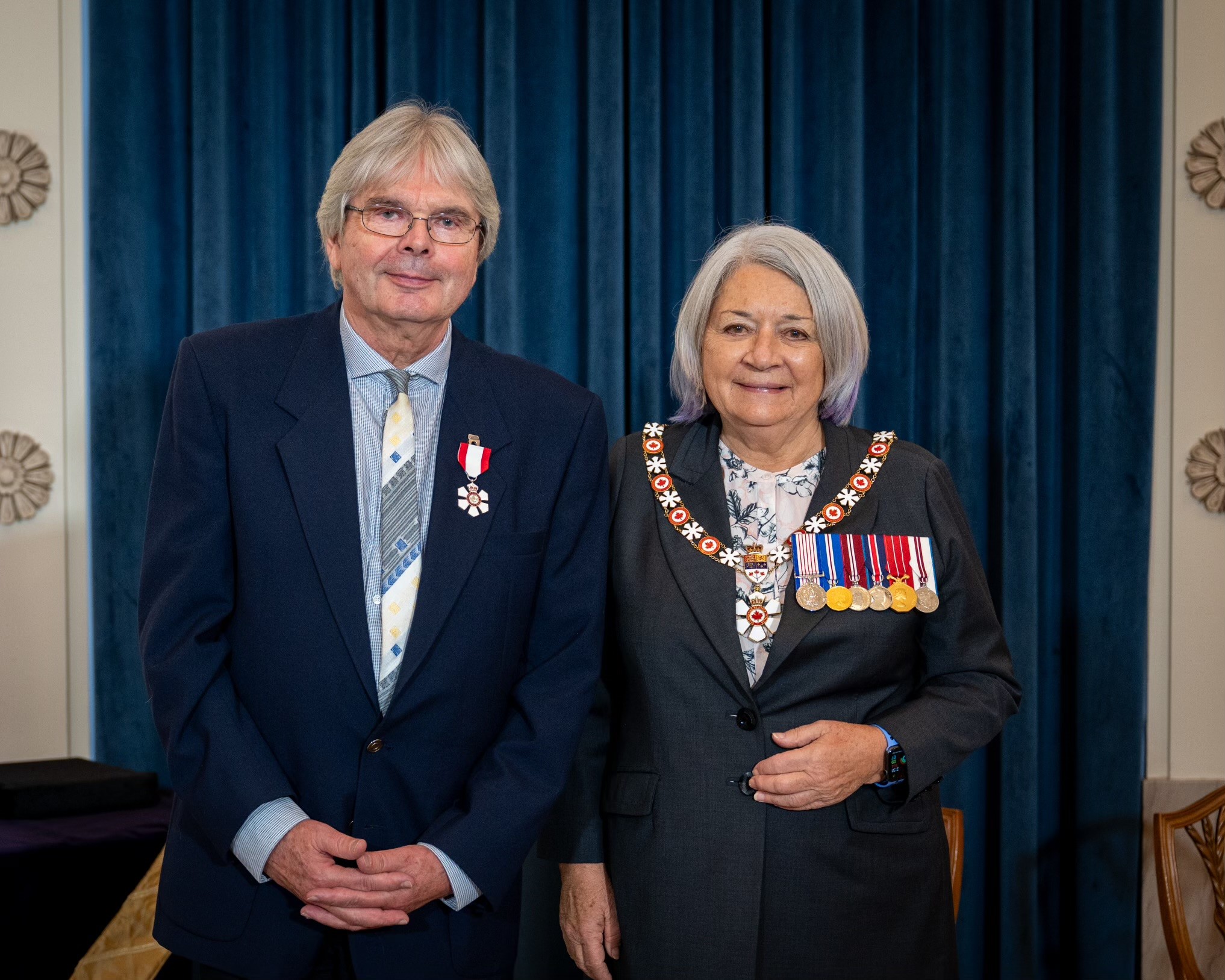 Un homme aux cheveux blancs portant l'insigne de l'Ordre du Canada sourit à la caméra à côté de la gouverneure générale Mary Simon, qui porte ses décorations. Tous deux se tiennent devant un rideau bleu.